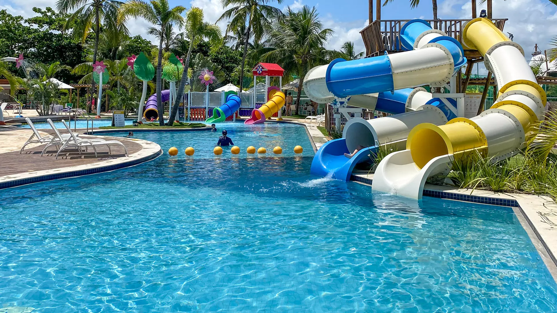 Piscina com toboáguas coloridos, cadeiras e palmeiras em resort à beira da praia durante o dia.